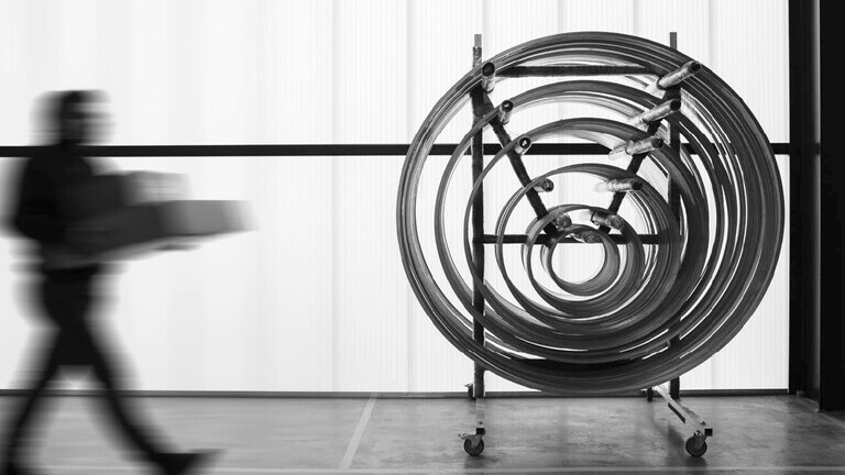 A black-and-white image shows a variety of rings of different diameters on a stand in SATTLER’s production hall; a person enters the frame from the left carrying a cardboard box. | © SATTLER