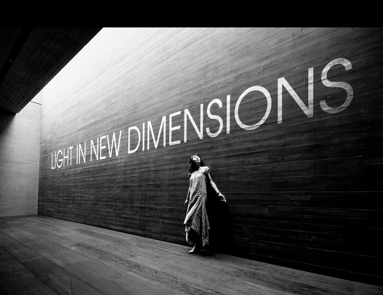 Schwarz weiß Bild mit einer Frau in einem leichten Kleid, die vor einer Betonwand steht  und von Licht angeleuchtet wird, ein Spiel von Licht und Schatten und dem Claim von SATTLER darauf: LIGHT IN NEW DIMENSIONS.