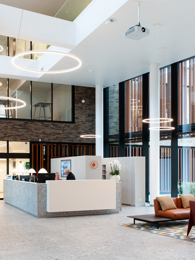Großzügiges Atrium mit Empfangstheke, Lounge und Klinkerwand, darüber schweben DOPPIO Ringleuchten von SATTLER in verschiedenen Größen und Höhen. | © HN