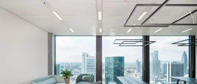 Lounge- und Meetingbereich mit Sofas und Holztischen vor bodentiefen Fenstern mit Blick auf die Frankfurter Skyline, darüber schwebende GIOCO Leuchten von SATTLER. | © © Kristof Lemp