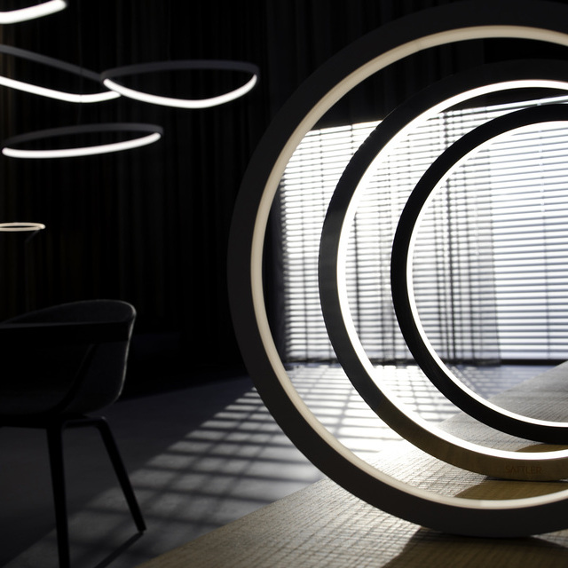 Dark photograph, the outline of a chair on the left, above it large pendant lights in various geometric shapes, in the foreground a wooden sideboard with 3 SLIM Table ring lights, which are offset and shine inwards. 