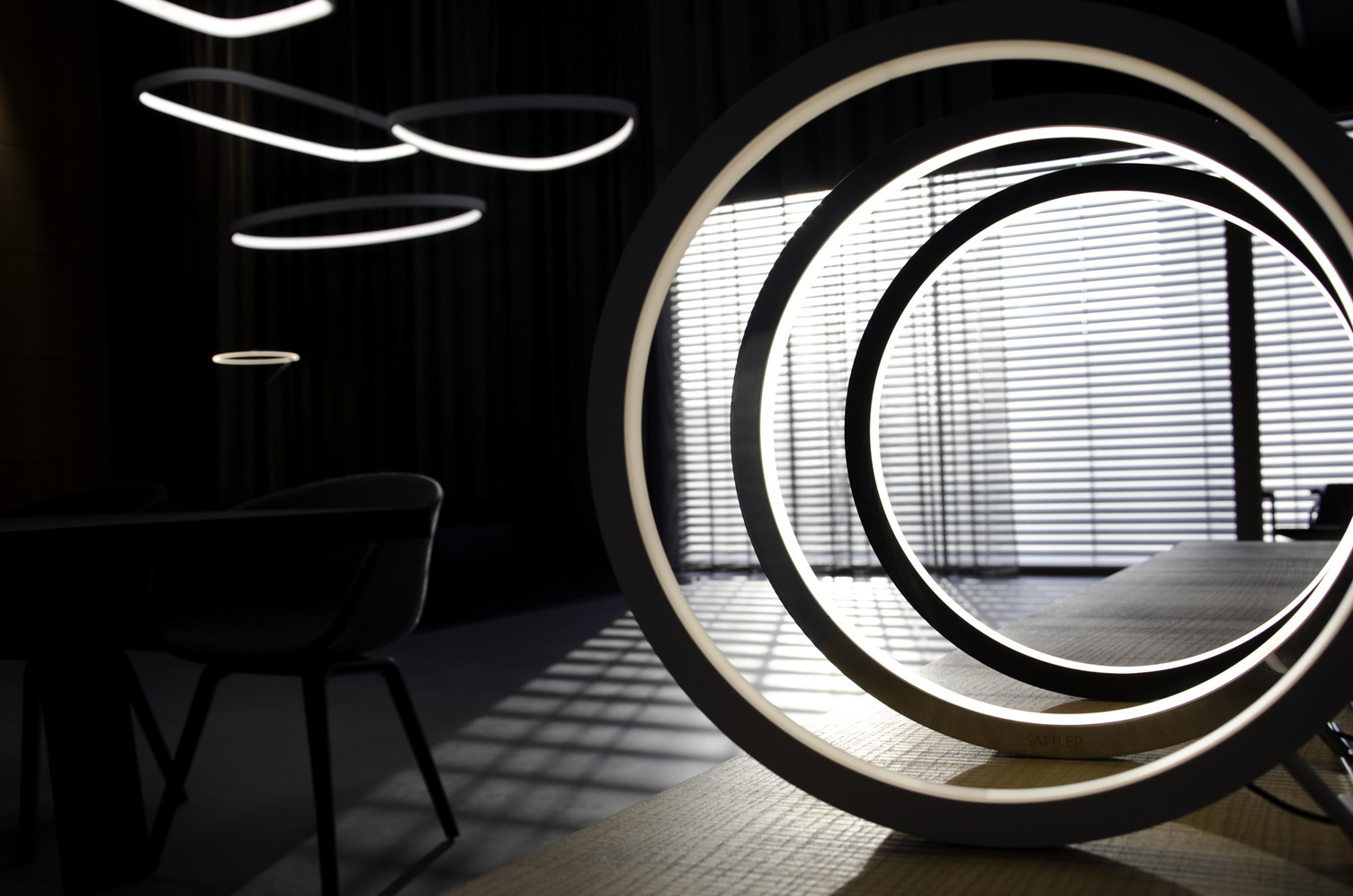 Dark photograph, the outline of a chair on the left, above it large pendant lights in various geometric shapes, in the foreground a wooden sideboard with 3 SLIM Table ring lights, which are offset and shine inwards.
