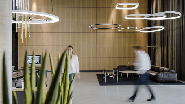 The image shows SATTLER’s large, open-plan showroom featuring several large pendant lights – Anello, Tia and Fio – along with various ring-shaped lights from the Soft series, a meeting table with a screen on one side, a leather sofa on the other side of the room, and two people walking across the room in different directions.