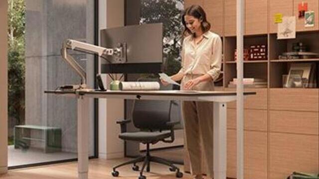 A woman is standing at a height-adjustable office workstation; to her left stands a tall floor lamp with a light panel, the Lavigo Single, by Waldmann. | © Waldmann