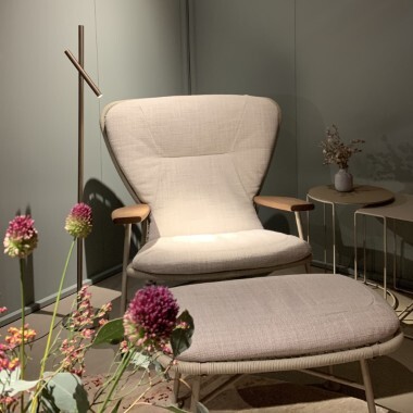 Blick über einen lila, grünen Blumenstrauß auf einen von einer zarten zylindrischen Bodenlampe, Fino Floor von Sattler, hell erleuchteten Loungesessel Shinzo von Walter Knoll, der vor einer grünen Wand steht auf der ein schwarzes Logo von Walter Knoll angebracht ist.
