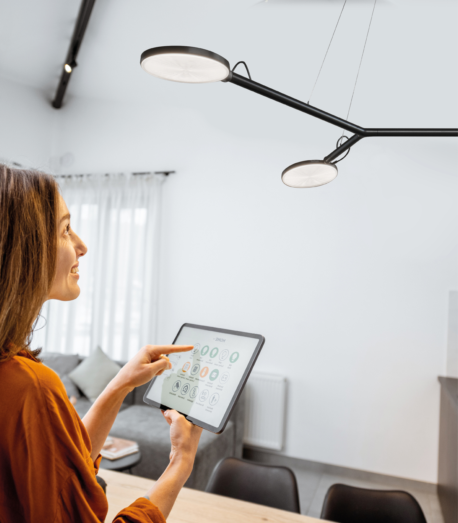 Eine junge lächhelnde Frau steht in einem Wohnzimmer mit einem Tablet in der Hand vor einer schwarzen AVVENI Pendelleuchte, grinst diese an und steuert diese per Fingerklick auf den Bildschirm.