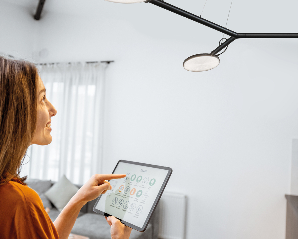 Smarte Steuerung über CASAMBI Eine junge lächhelnde Frau steht in einem Wohnzimmer mit einem Tablet in der Hand vor einer schwarzen AVVENI Pendelleuchte, grinst diese an und steuert diese per Fingerklick auf den Bildschirm.