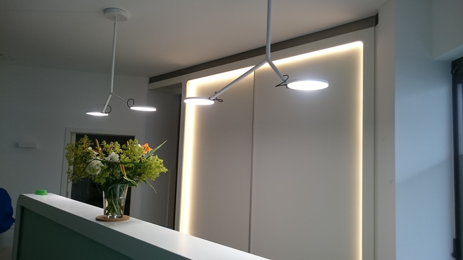 Weiße Empfangstheke mit zwei weißen einfach verzweigten Anbauleuchten AVVENI CEILING 2. Jede Leuchte endet in zwei beweglichen runden flachen Leuchtenköpfe.