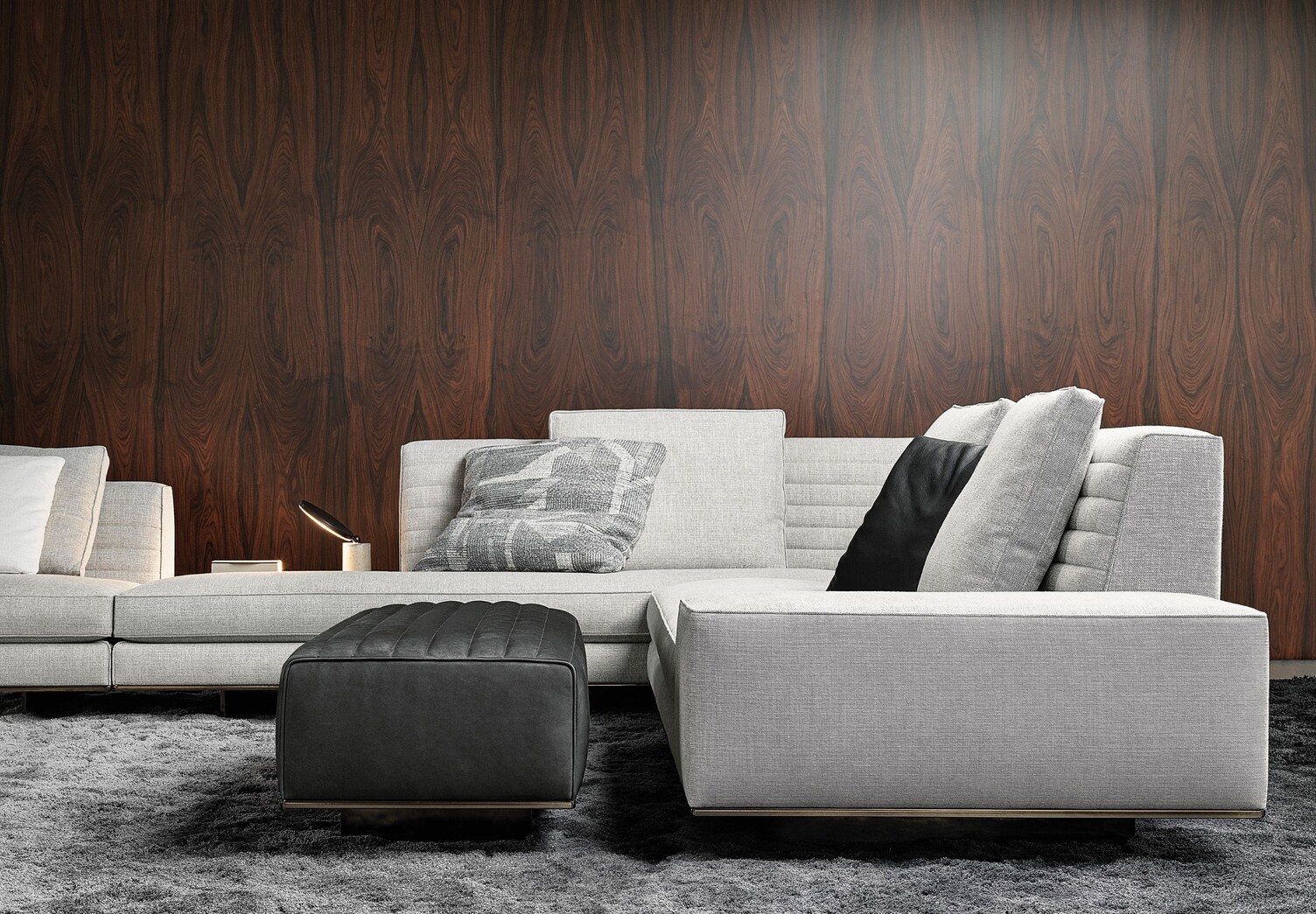 Hellgraue elegante Eck-Sofalandschaft von Minotti vor einer dunklen edlen Holzwand. Wandnah ist die Tischleuchte AVVENI Concrete bestehend aus einem hellem Betonsockel und einem runden flachen schwarzen Metallleuchtenkopf leuchtend zu sehen. 