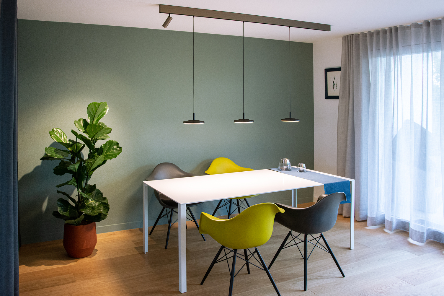 Lichtelement Pendelleuchte AVVENI PENDANT 1 für PALADIO TRACK Modernes Esszimmer mit weißem rechteckigen Tisch und 4 bunten Stühlen darum. Über dem Tisch ist eine Stromschiene an die Decke angebaut, in dieser ist am linken Rand ein zylindrischer Spot (EVOLUTION SPOT S) eingesetzt, der die Pflanze im linken Zimmer Eck beleuchtete, und 3 schwarze Pendelleuchten mit runden flachen Leuchtenköpfen (AVVENI PENDANT 1), die den Tisch ausleuchten.