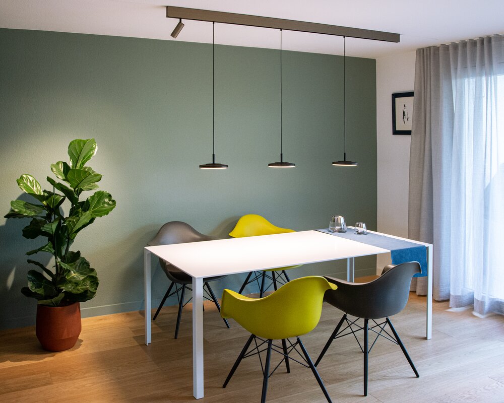 Lichtelement Pendelleuchte AVVENI PENDANT 1 für PALADIO TRACK Modernes Esszimmer mit weißem rechteckigen Tisch und 4 bunten Stühlen darum. Über dem Tisch ist eine Stromschiene an die Decke angebaut, in dieser ist am linken Rand ein zylindrischer Spot (EVOLUTION SPOT S) eingesetzt, der die Pflanze im linken Zimmer Eck beleuchtete, und 3 schwarze Pendelleuchten mit runden flachen Leuchtenköpfen (AVVENI PENDANT 1), die den Tisch ausleuchten.