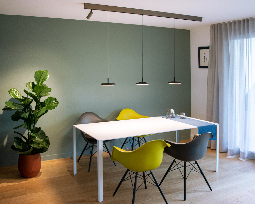 Lichtelement Pendelleuchte AVVENI PENDANT 1 für PALADIO TRACK Modernes Esszimmer mit weißem rechteckigen Tisch und 4 bunten Stühlen darum. Über dem Tisch ist eine Stromschiene an die Decke angebaut, in dieser ist am linken Rand ein zylindrischer Spot (EVOLUTION SPOT S) eingesetzt, der die Pflanze im linken Zimmer Eck beleuchtete, und 3 schwarze Pendelleuchten mit runden flachen Leuchtenköpfen (AVVENI PENDANT 1), die den Tisch ausleuchten.