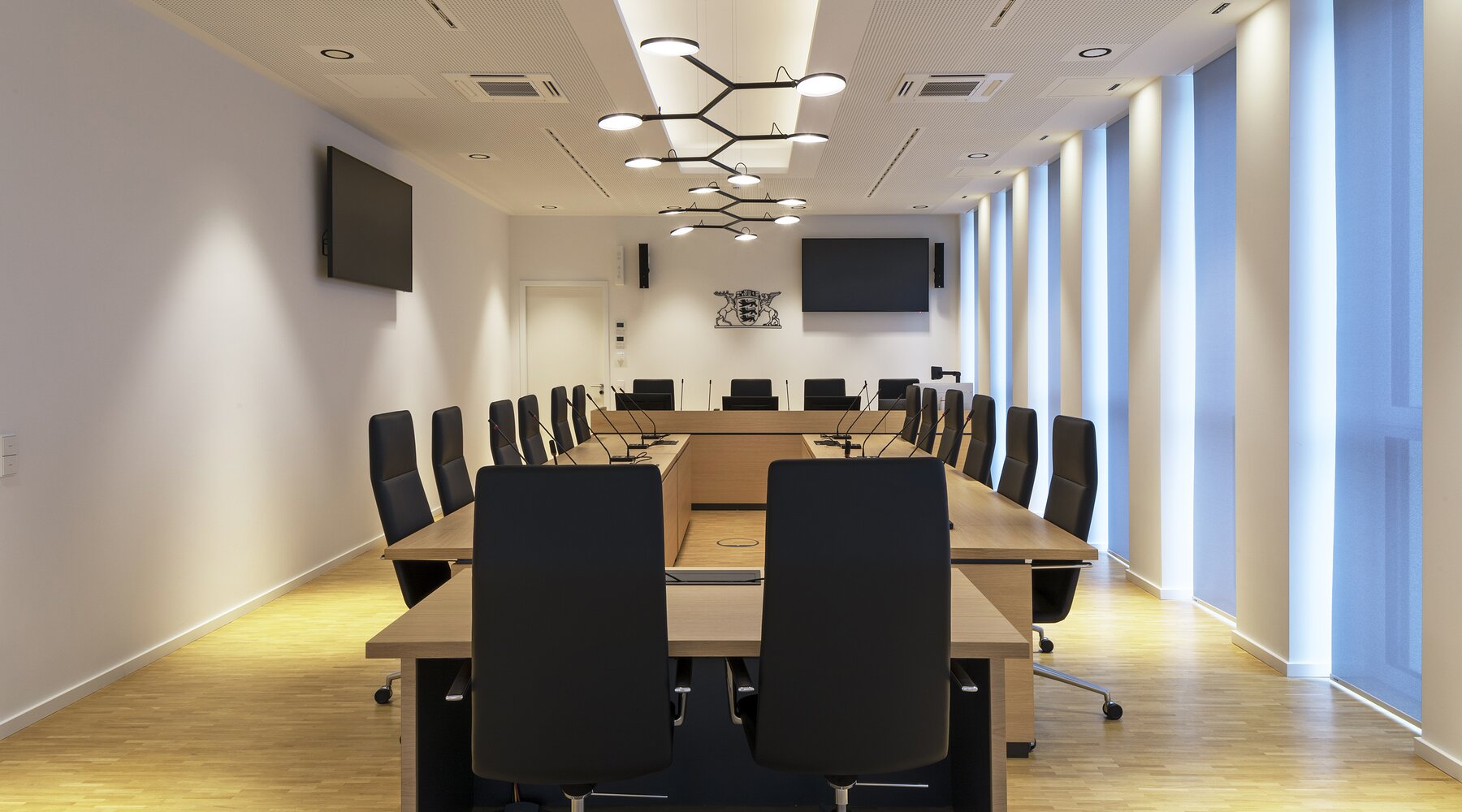 Two linear branched pendant luminaires with flat light heads, AVVENI Pendant 6, Stuttgart Fasanenhof District Court: courtroom in wood look with black chairs and the Baden Württemberg logo in the centre. | © Juliane Eirich