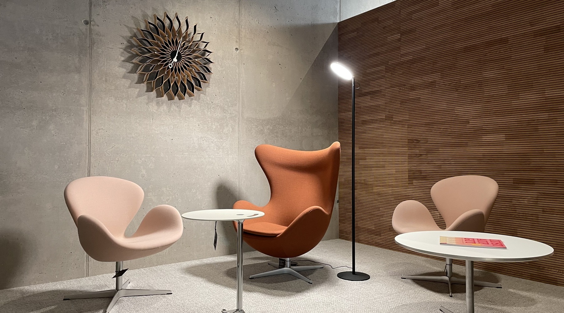 Stehleuchte mit einem flachen, runden Leuchtenkopf (AVVENI Floor) in einem Loungebereich mit drei Sesseln in rötlichen Erdtönen der Firma STREIT und einer modernen verspielten Wanduhr vor einer Sichtbetonwand. | © STREIT