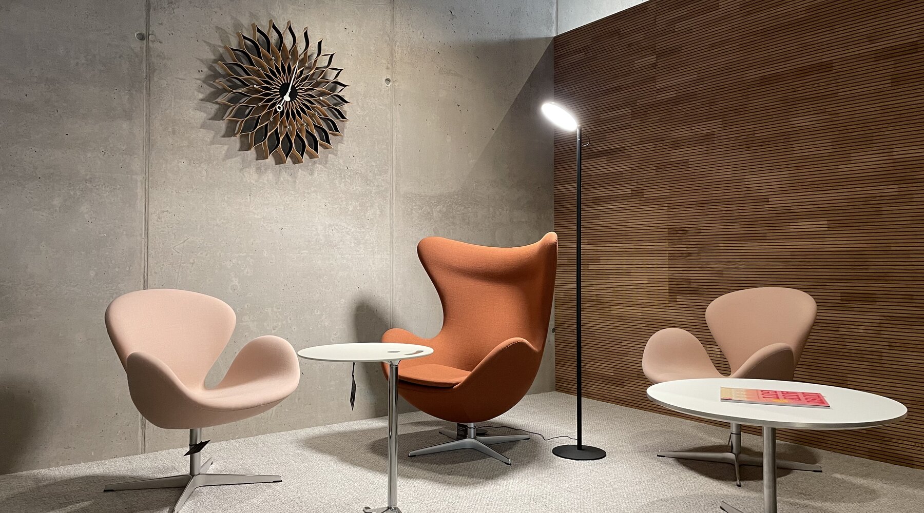 Stehleuchte mit einem flachen, runden Leuchtenkopf (AVVENI Floor) in einem Loungebereich mit drei Sesseln in rötlichen Erdtönen der Firma STREIT und einer modernen verspielten Wanduhr vor einer Sichtbetonwand. | © STREIT