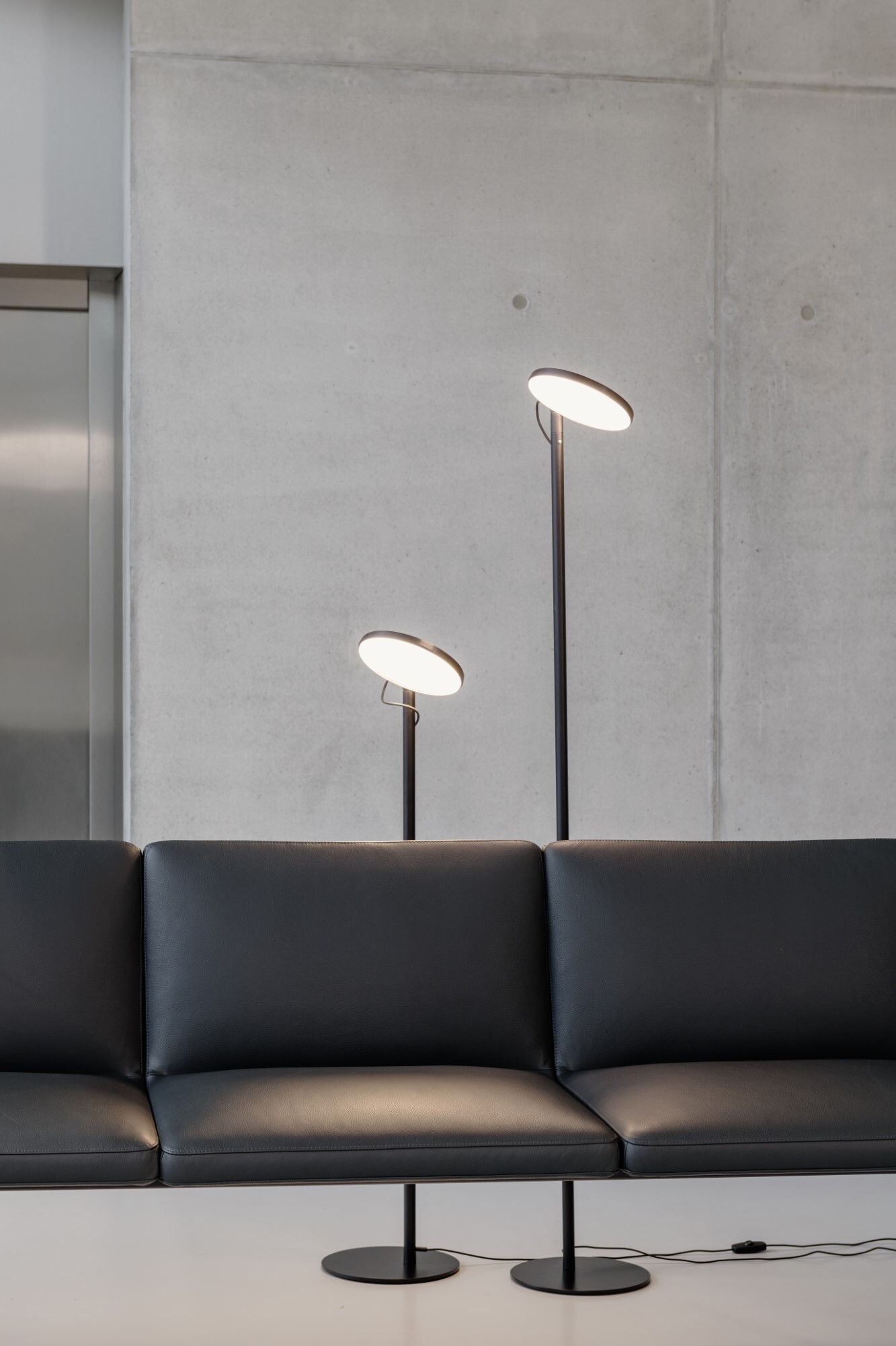 Lounge Brunner Group - AVVENI FLOOR | © bildhübsche fotografie | Andreas Körner Zwei schwarze Stehleuchten der Serie AVVENI FlOOR unterschiedlicher Höhe mit rundem flachen Leuchtenkopf hinter schwarzem Business Ledersofa der Brunner Group. | © bildhübsche fotografie | Andreas Körner