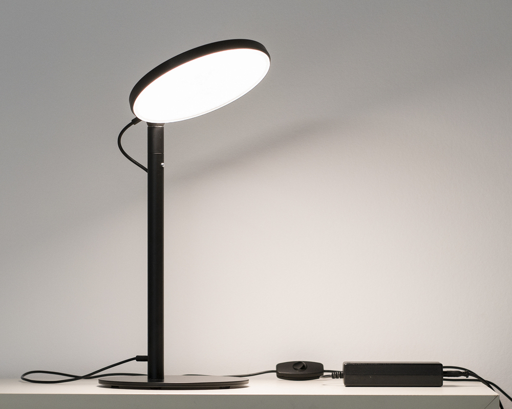 Black table lamp with power supply + switch Black table lamp with flat, round, plate-shaped head and dimmer button on the base of the lamp, power supply unit separately adjacent.