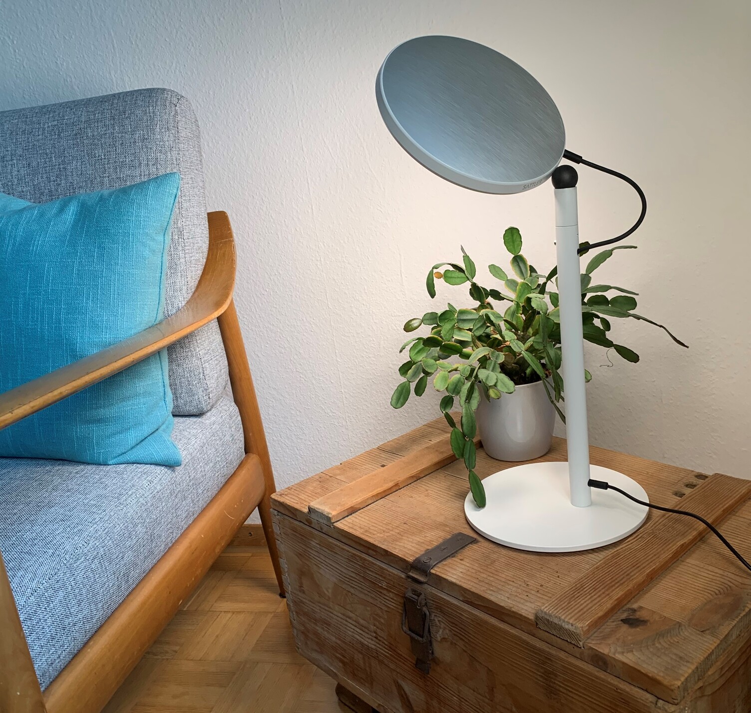 Table lamp AVVENI TABLE in the reading corner Avveni Table floor lamp with plate-shaped silver aluminium lamp head and white round lamp base on a wooden chest with plant and 60s armchair with blue cushion.