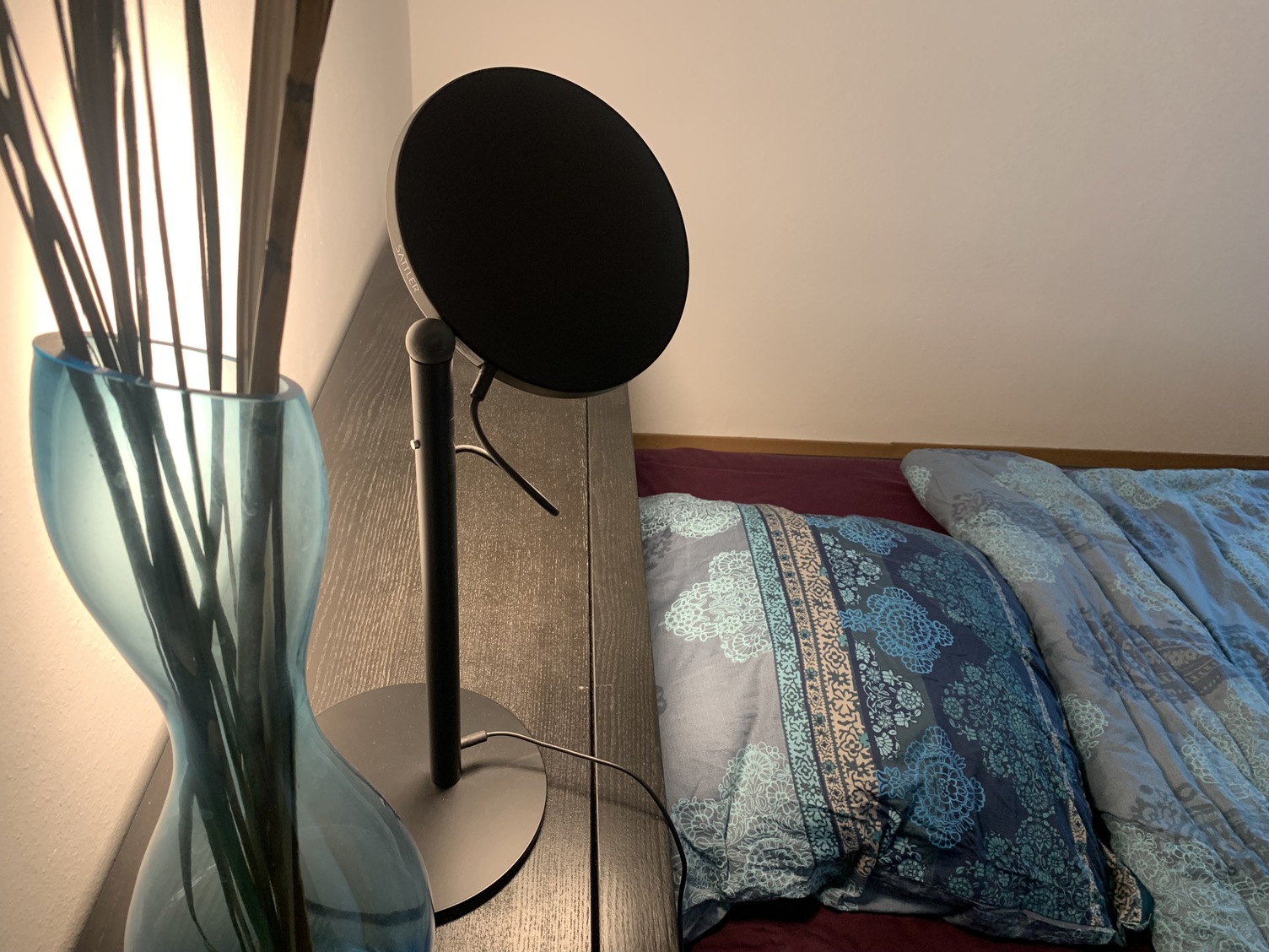 Reading Light - AVVENI TABLE in the bedroom Avveni Table lamp with plate-shaped black aluminium lamp head and black round lamp base on the deep headboard of a black wooden bed with blue bed linen.