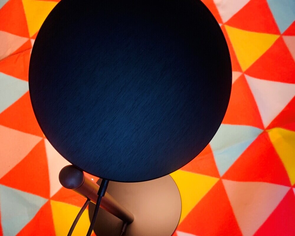 Surface AVVENI light head top cover: brushed and anodised View of the head of a black table lamp consisting of a round, flat lamp head and a cylindrical base, standing on a colourful picnic blanket.