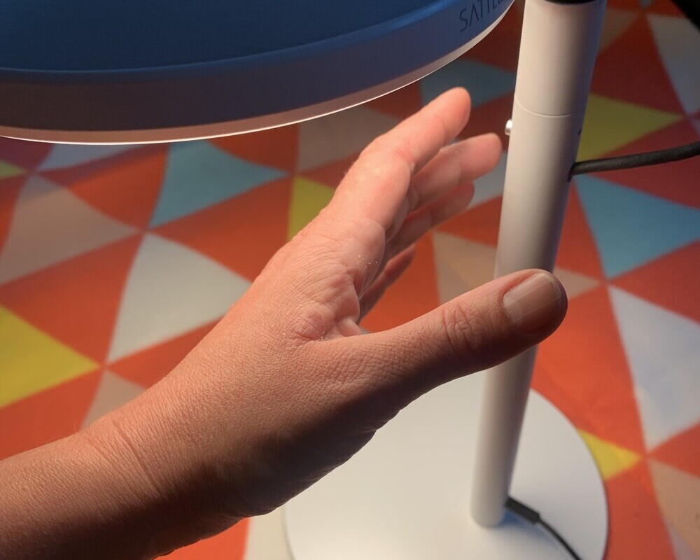 Round push-button dimmer Hand on the dimmer button in the light stand of the plate-shaped AVVENI TABLE lamp in silver and white on a colourful graphic ceiling.