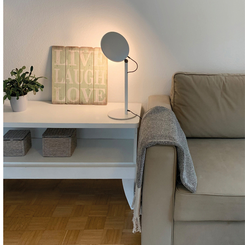 Light for Live, Laugh, Love. White sideboard with 2 rectangular baskets, on which stands a plant and a green sign reading ‘Live, Laugh, Love’, illuminated by a white floor lamp, AVVENI TABLE, with movable round lamp head. Next to it is a beige sofa with a woollen blanket over the backrest.