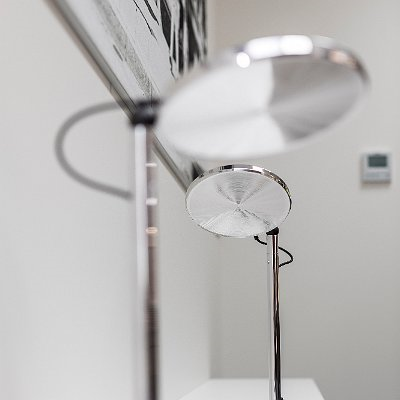 Circular prismatic diffuser Two table lamps with round, flat lamp heads that are switched off and show the circular prismatic diffuser. The lamp body is a cylindrical base made of polished aluminium.