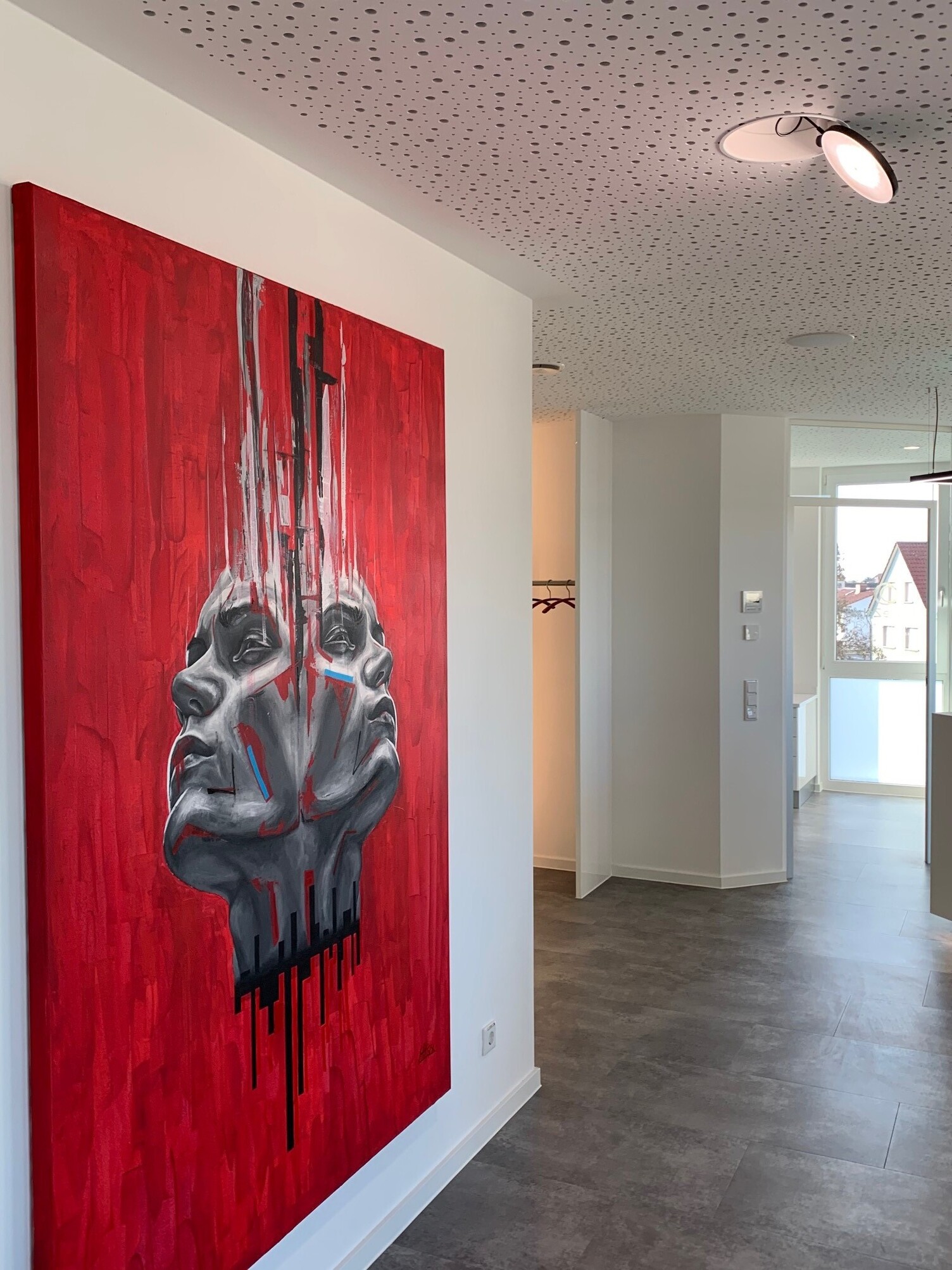 Wandbildbeleuchtung mit dreh- und klappbarem AVVENI Downlight (Deckeneinbau mit Rand) Weißer Flur. An der linken Wand hängt ein großes gemaltes Bild mit zwei Köpfen auf roter Leinwand. Das Bild wird von einem Strahler in der weißen Lochdecke, dem AVVENI DOWNLIGHT with frame, beleuchtet.