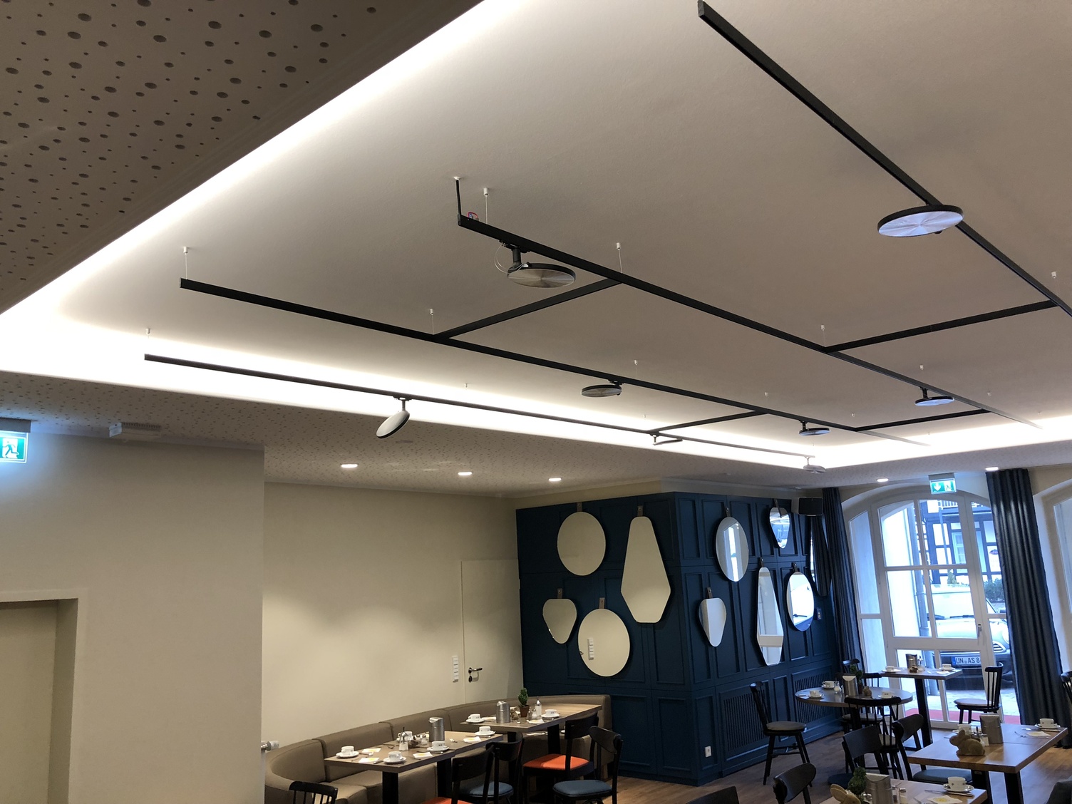 Suspended track lighting system lightlight + AVVENI Focus on the ceiling with black, connected, branched, flexible light track system lightlight from buschfeld with AVVENI lighting heads on the ceiling above tables in hotel restaurant.