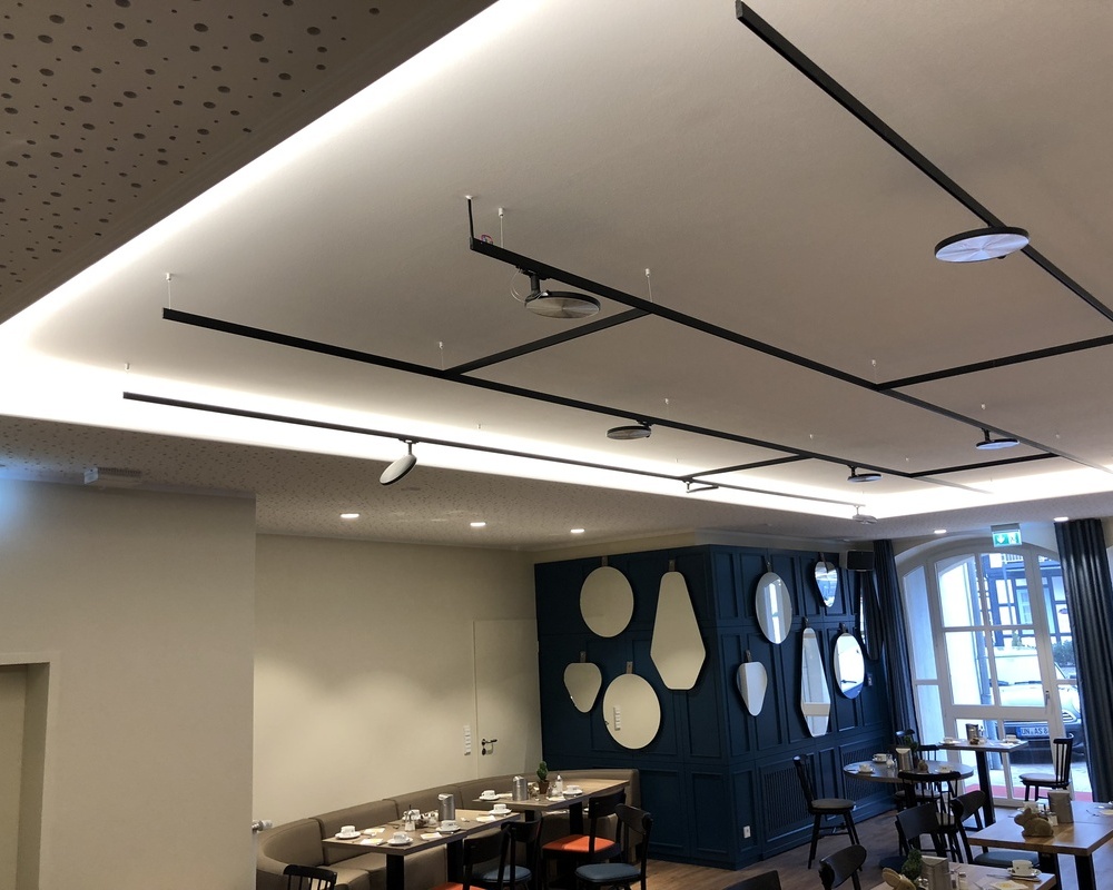 Suspended track lighting system lightlight + AVVENI Focus on the ceiling with black, connected, branched, flexible light track system lightlight from buschfeld with AVVENI lighting heads on the ceiling above tables in hotel restaurant.