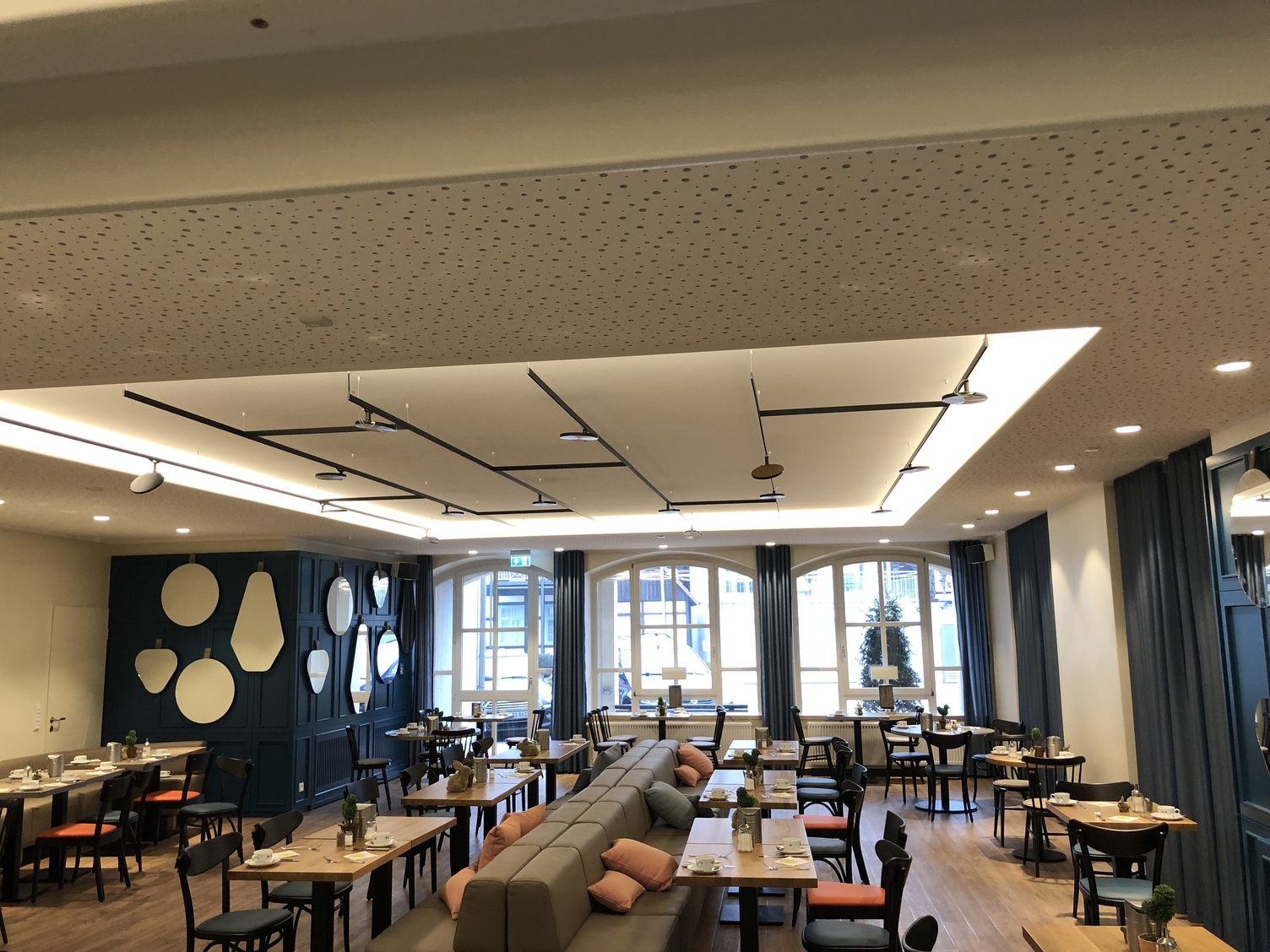 Suspended track lighting system in hotel restaurant Black, connected, branched, flexible light track system lightlight from buschfeld with AVVENI lighting heads on the ceiling above tables in a hotel restaurant.