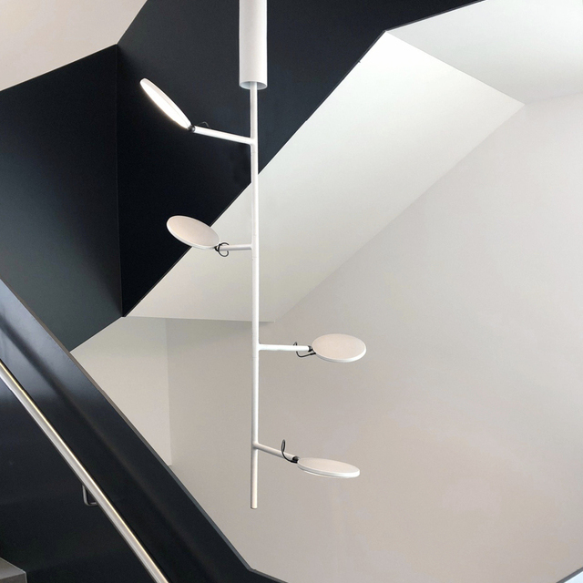 Modernes Treppenhaus mit Geländer aus schwarzen Metallwangen, in der Mitte des Treppenauges die empfohlene Lampe fürs Treppenhaus: Hängelampe AVVENI VERTICAL in weiß. Ein auf die Länge verzweigter  runder Stab an dessen Ästen runde flache Leuchtenköpfe positioniert sind, die in verschiedene Raumrichtungen gedreht werden können. Licht auf Treppenstufe oder Wand. | © Garmo Stuttgart