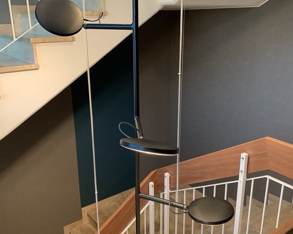 Bewegliche Leuchtenköpfe zur Treppenstufenbeleuchtung Blick von der Seite auf: Drei schwarze Pendelleuchten in einem Treppenauge bestehend aus je einem Rohr von dem umlaufend und höhenversetzt 6 bewegliche Leuchtenköpfe an kleinen Armen weggehen. Die AVVENI Pendant 6, in unterschiedlichen Höhen wirken wie stilistische Blumen im gefliesten, geraden mehrläufigen Treppenhaus des Kurparkhotel Bad Wilhelmshöhe.