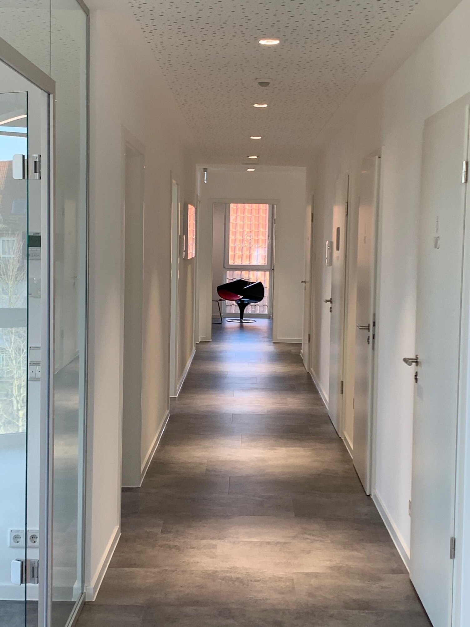 Corridor lighting with downlights BE3 round A long corridor leading to several doors. White walls and a white perforated ceiling. Round BE3 downlights are distributed along the ceiling, casting pools of light onto the corridor.