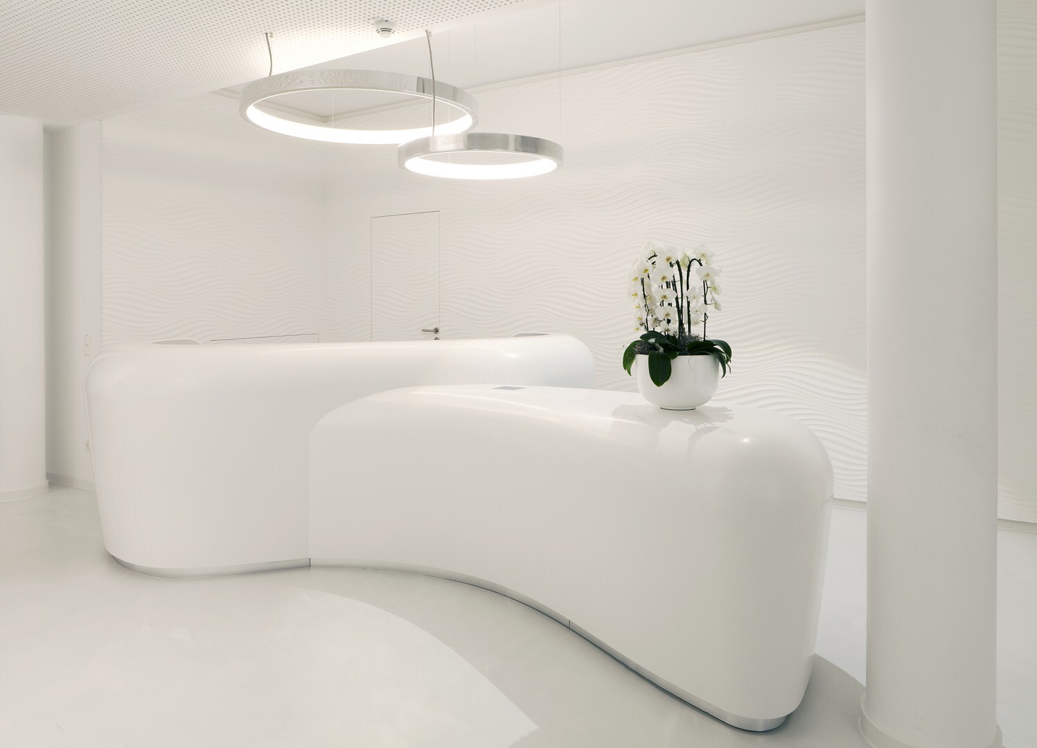 Weißer Raum mit zwei weißen futuristischen Empfangstheken mit Orchideen auf einer, über den Theken sind 2 Aluminium Ringleuchten CIRCOLO abgependelt sind.