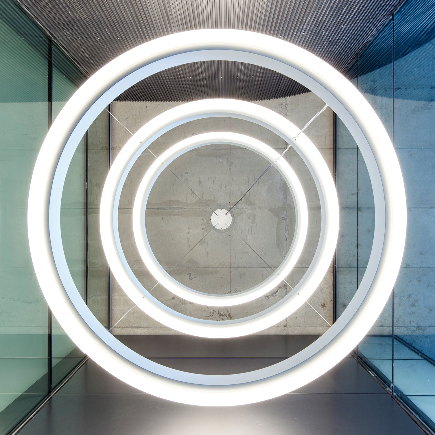 Ansicht von unten auf drei nach außen strahlende große Ringleuchten, CIRCOLO INVERSE mit 1,32 m Durchmesser, die untereinander in einem Glasturm, in Frankfurt Westhafen - Pier Relaunch, abgehangen sind.