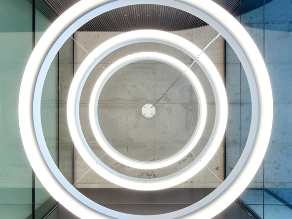Ansicht von unten auf drei nach außen strahlende große Ringleuchten, CIRCOLO INVERSE mit 1,32 m Durchmesser, die untereinander in einem Glasturm, in Frankfurt Westhafen - Pier Relaunch, abgehangen sind. | © Barbara Burg | Oliver Schuh, Palladium Photodesign, Köln