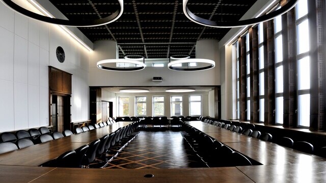 Großer Sitzungssaal der IHK Duisburg mit dunklen Möbeln, Holztisch in U-Form angeordnet, mit mehreren großen Ringleuchten (CIRCOLO & CIRCOLO INVERSE.) im Durchmesser 2,6 m, die von der Decke abgehängt oder angebaut sind.  | © Wibke Evert