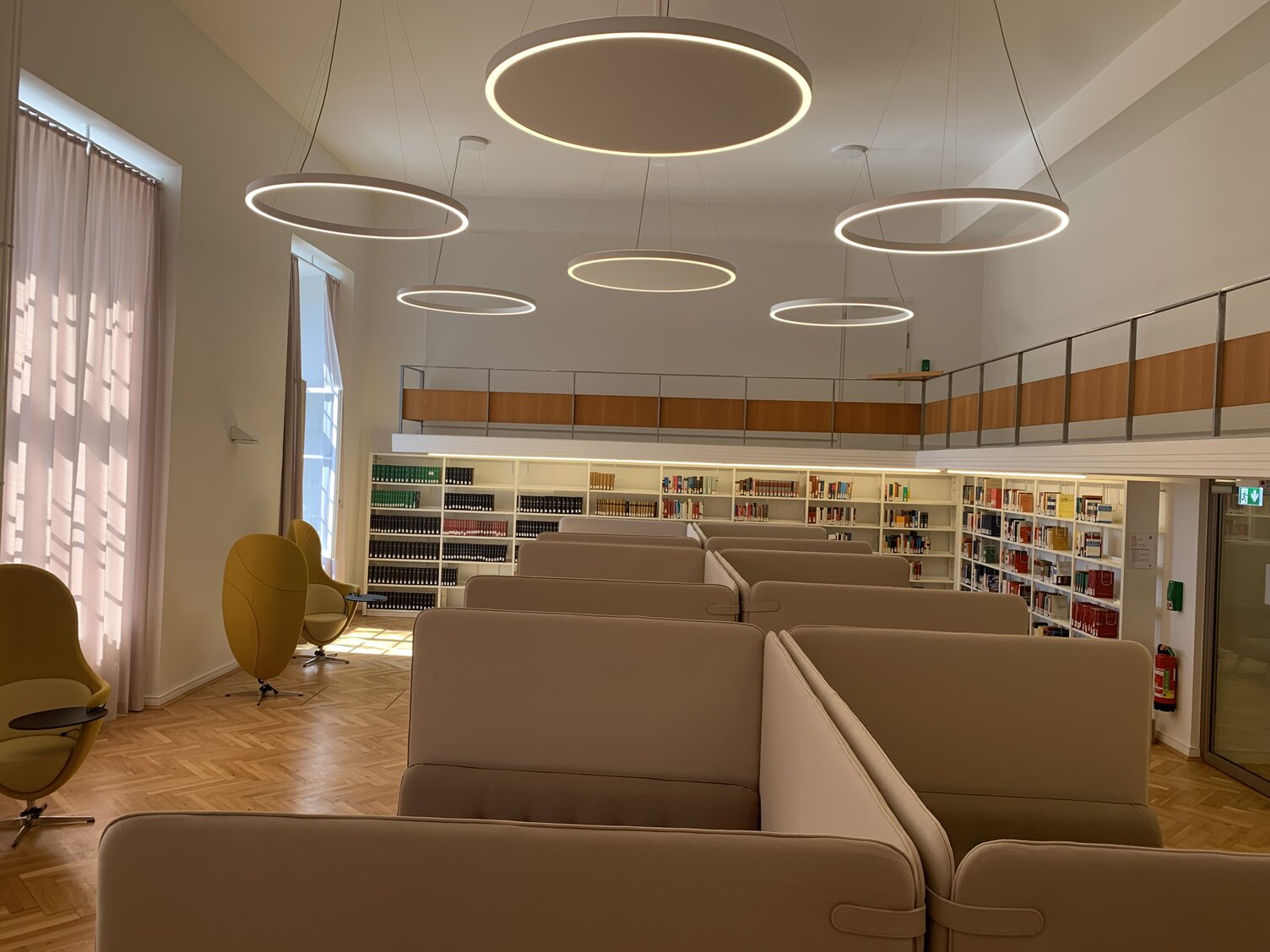 Bibliothek der TU Schloß Darmstadt mit DUETTO Ringleuchten Bibliothek der Technischen Universität Darmstadt mit Bücherregalen an der Wand, in der Mitte des Raumes Akustikbänke von der Decke sind mehrere DUETTO Ringleuchten abgehangen, teilweise mit akustischwirksamen Stoff in der Leuchtenmitte.