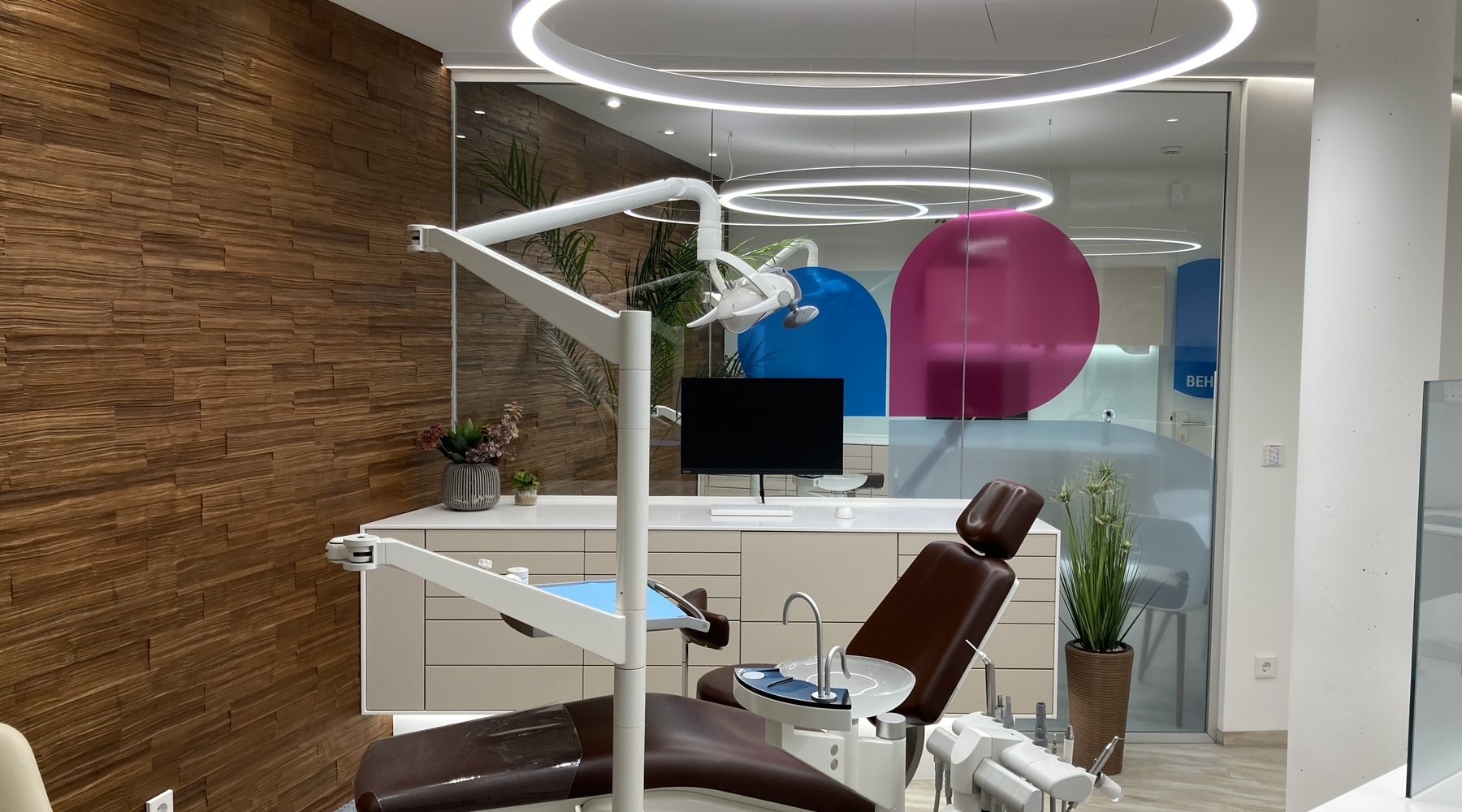 Optional: LED High Output Zahnarztstuhl vor einer modernen braunen Strukturwand, darüber eine runde Ringleuchte mit hohem Lumenoutput (DUETTO MED).