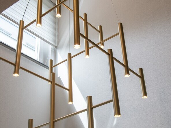 Moderner Kronleuchter für Treppenauge eines Privathauses: EVOLUTION VERTICAL 1. Großansicht des tubusfömigen goldenen Kronleuchters zwischen vier weißen Wänden, eine Pendelleuchte für ein Treppenauge eines modernen Wohnhauses. | © SATTLER