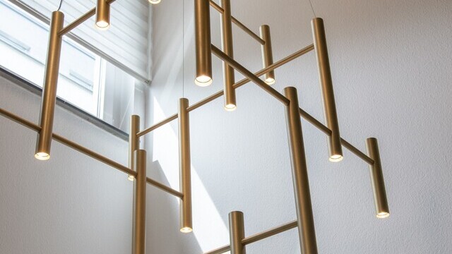 Modern chandelier for staircase eye: EVOLUTION VERTICAL 1: Zoom to a tubular golden chandelier, shown between 4 white walls, a pendant lamp for a staircase eye of a private residence. | © SATTLER