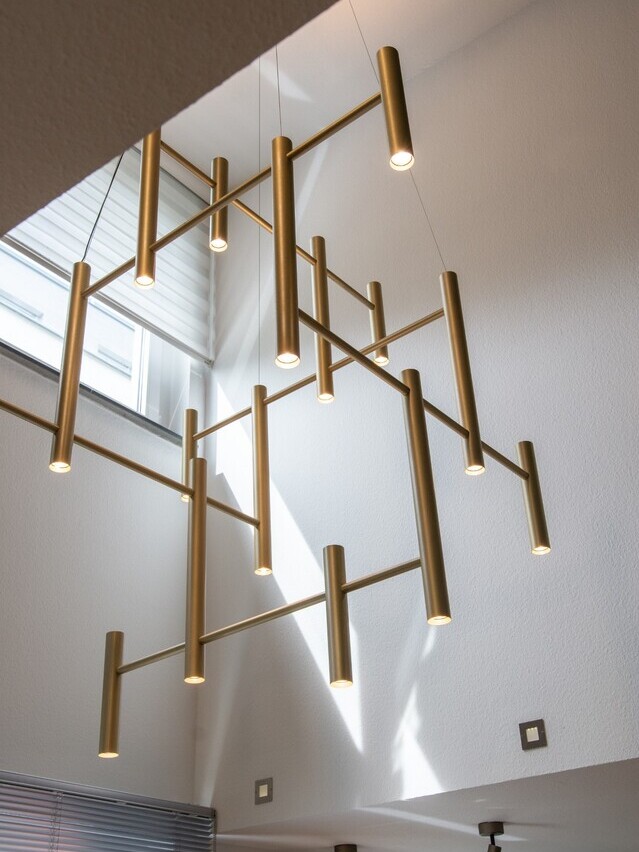 Moderner Kronleuchter für Treppenauge eines Privathauses: EVOLUTION VERTICAL 1. Großansicht des tubusfömigen goldenen Kronleuchters zwischen vier weißen Wänden, eine Pendelleuchte für ein Treppenauge eines modernen Wohnhauses. | © SATTLER
