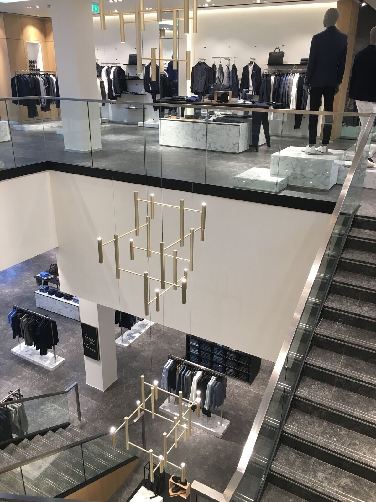 View of a pendant luminaire consisting of three modern golden chandeliers (EVOLUTION VERTICAL 3), in a stone staircase with glass stringers of a textile shop for modern, elegant menswear.