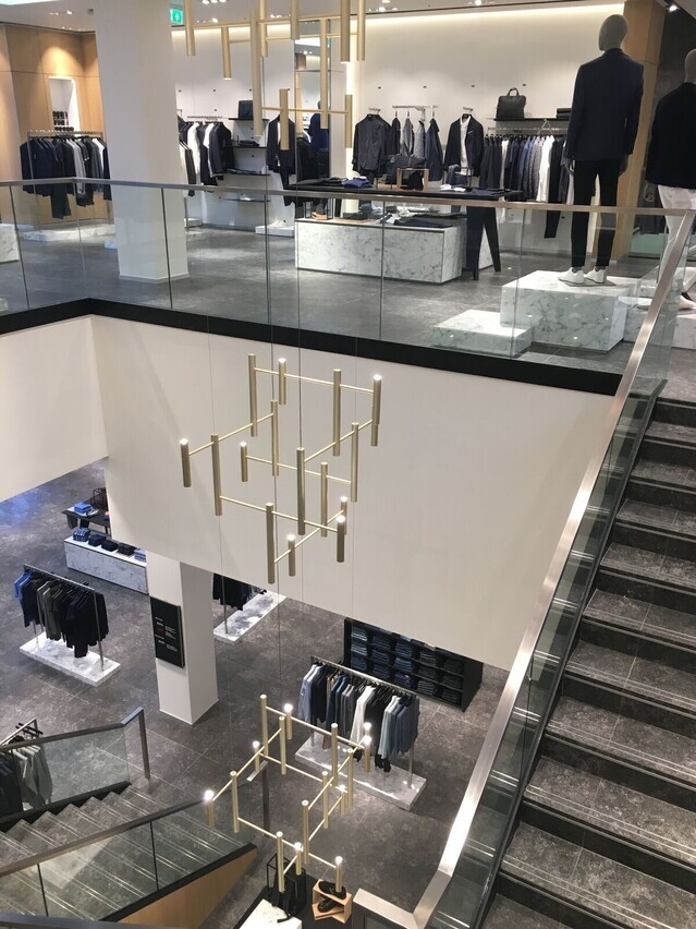 Blick auf eine Pendelleuchte bestehend aus drei modernen goldenen Kronleuchtern (EVOLUTION VERTICAL 3), in einem Steintreppenhauses mit Glaswangen eines Textilgeschäftes für moderne, edle Herrenbekleidung.
