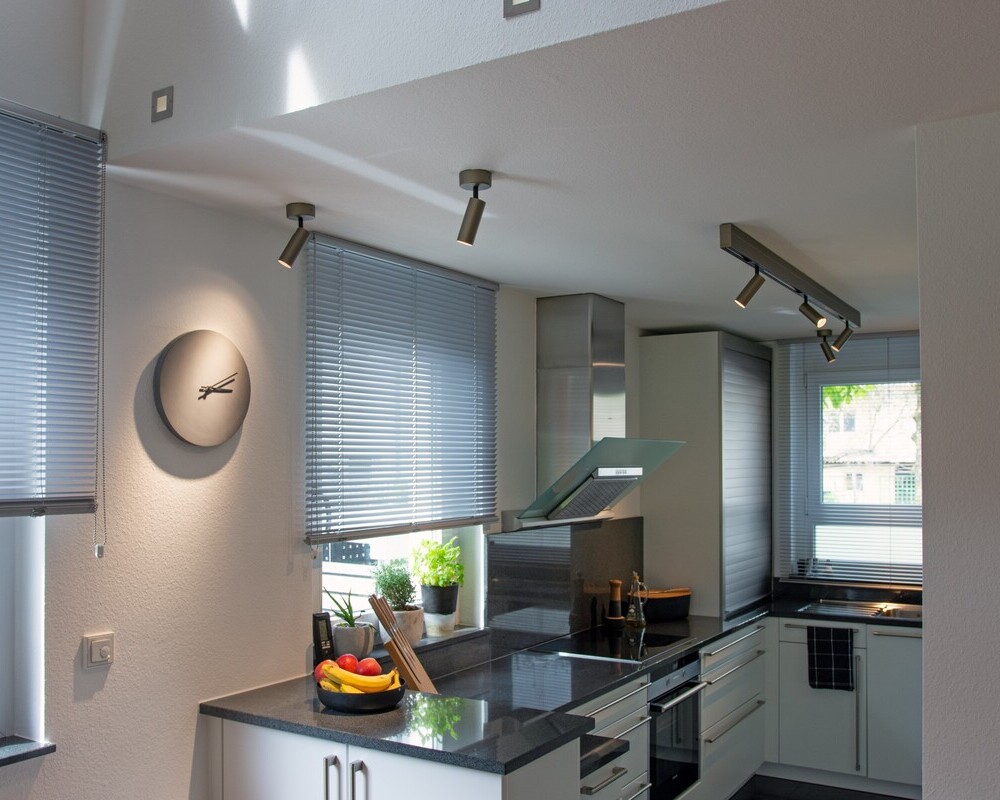 Surface-mounted luminaires: adjustable EVOLUTION SPOT & flexible PALADIO TRACK Open-plan kitchen unit with two cylindrical EVOLUTION SPOT spotlights, mounted directly to the ceiling via the ceiling canopy; one illuminates the kitchen clock. Along the kitchen unit, a linear surface-mounted luminaire, PALADIO TRACK, into which 3 EVOLUTION SPOTs are clicked, each directed in different beam angles to illuminate the entire kitchen worktop.