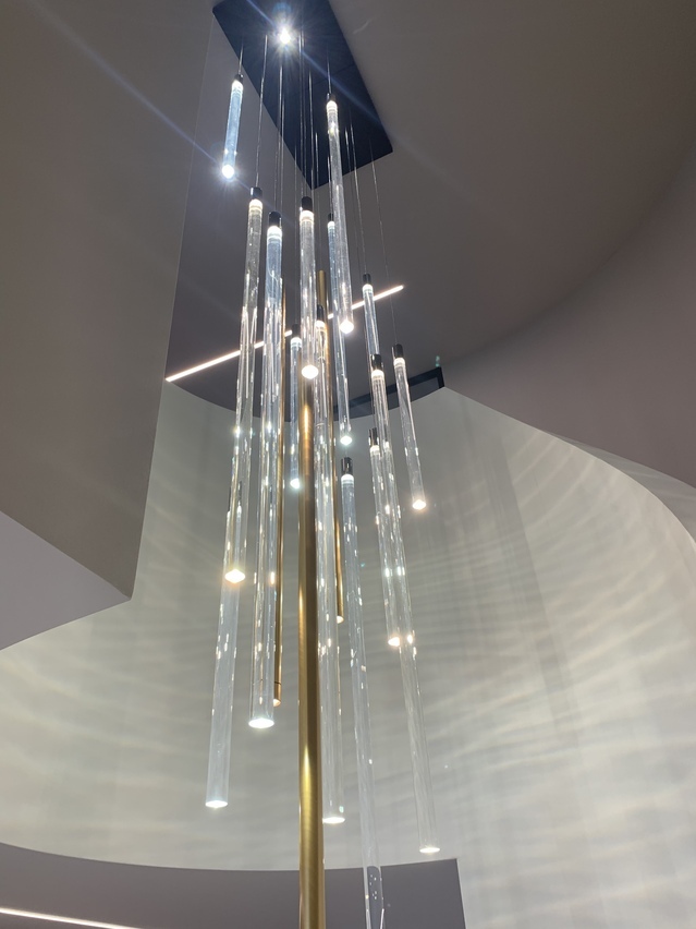 Blick auf ein Leuchtenensamble der zylindrischen Pendelleuchten FIO, aus runden, transparenten Acrylstäben, und TIA, aus goldenem Aluminiumtubus, in einem Treppenauge einer Betontreppe.