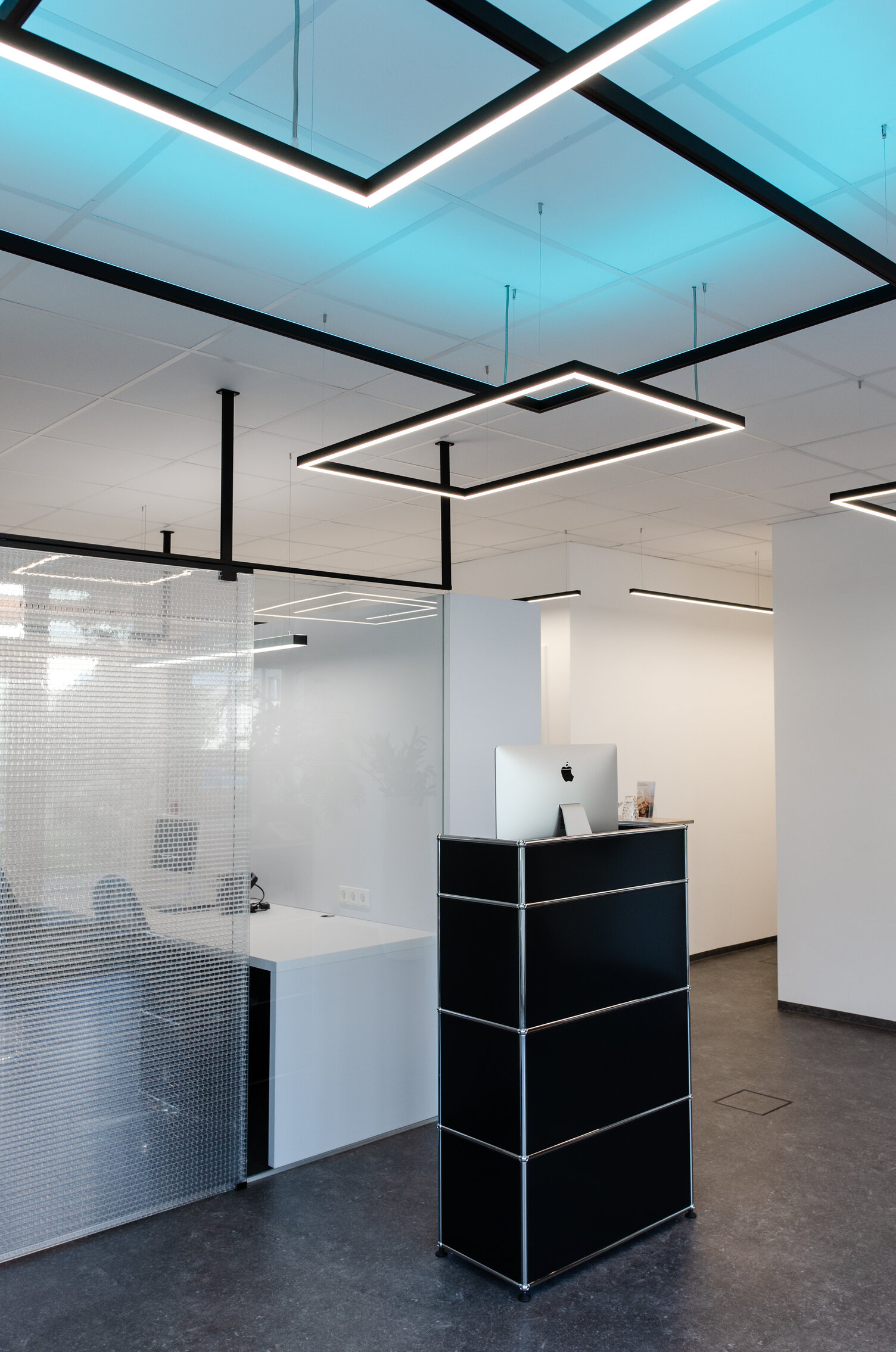 Kleiner schwarzer Empfangstresen vor einem Büro, das mit einer Glaswand abgetrennt ist, weiße Wände und weiße Decke, von der viereckige Pendelleuchten der GIOCO Serie abgehangen sind, zwei leuchten direkt weiß und eine indirekt in RGB Blau gegen die Decke.
