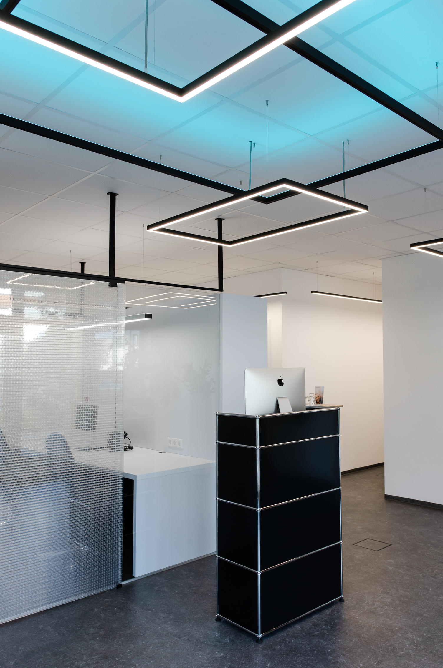 Small black reception desk in front of an office separated by a glass wall, white walls and white ceiling from which square pendant luminaires from the GIOCO series are suspended, two shine directly in white and one indirectly in RGB blue against the ceiling.