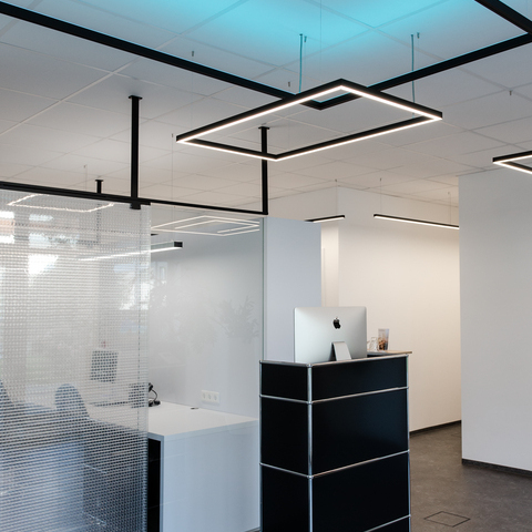 Small black reception desk in front of an office separated by a glass wall, white walls and white ceiling from which square pendant luminaires from the GIOCO series are suspended, two shine directly in white and one indirectly in RGB blue against the ceiling.